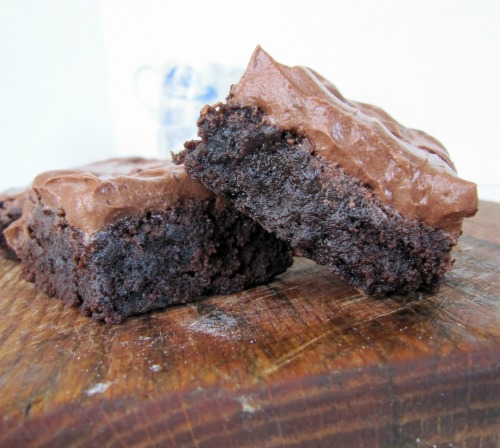 close up of two gooey fudgy cocoa brownies with chocolate buttercream frosting leaning against each other on a wooden cutting board.