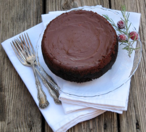 Overhead shot of a large round chocolate cheesecake on a clear glass platter on a stack of white cloth napkins. Two silver forks are to the side of the cheesecake which is resting on a dark wood brown table.