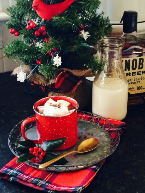 A red mug filled with rich spiced bourbon hot chocolate spiced with cinnamon and whiskey is on a round metal tray along with a serving spoon. The tray is in front of a glass bottle of milk and a bottle of bourbon alongside a Christmas tree.
