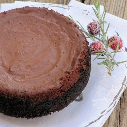Large chocolate cheesecake with Oreo crust on a clear glass platter with a sprig of cranberries as a garnish. The platter is on a white tablecloth