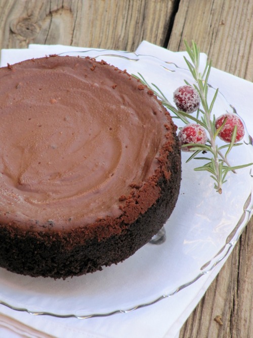 Large chocolate cheesecake with Oreo crust on a clear glass platter with a sprig of cranberries as a garnish. The platter is on a white tablecloth
