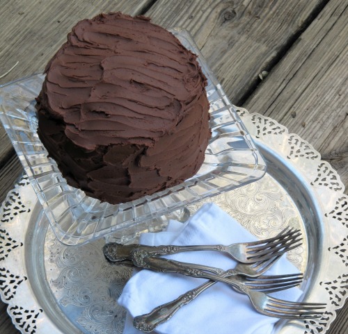 Overhead view of a round chocolate layer cake covered with dark chocolate fudge frosting, sitting on a square glass cake stand on a silver serving platter.