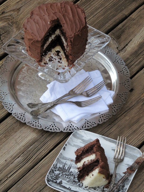 A large two layer chocolate cake with dark chocolate fudge frosting is sitting on a glass cake stand on a silver tray. A slice is taken out of the cake and is on a plate in front of the cake stand.