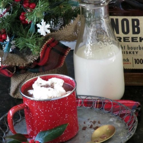 A ded enamel mug of bourbon spiced hot chocolate topped with whipped cream and chocolate shavings sits on a round metal tray in front of a glass bottle of milk and a bottle of bourbon and Christmas decorations