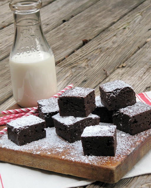 side view of fudgy chocolate brownies on a wooden cutting board next to red and white straws and a glass of milk.