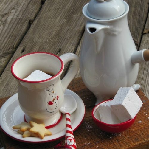 A tall cup of irish hot chocolate with marshmallow topping on a saucer with little sugar cookies next to a small bowl of homemade marshmallows in front of a tall white pitcher