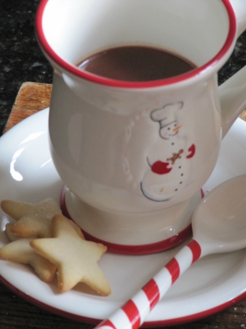 A tall red-rimmed white ceramic mug holds creamy Irish Hot Chocolate. The cup is on a matching plate along with some small star-shaped sugar cookies and a spoon with a red-and-white-striped candy cane handle.