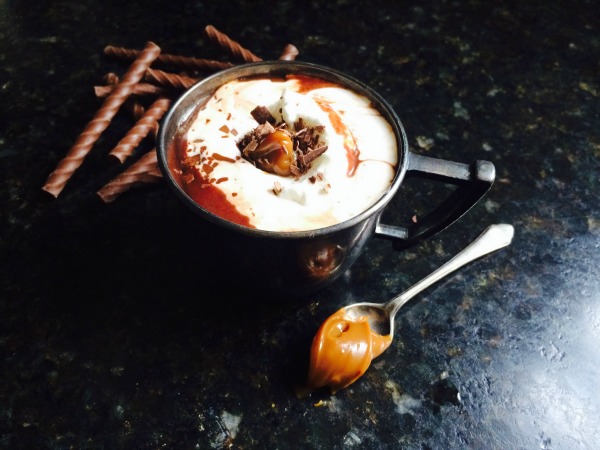 closeup overhead view of a dark gray mug ofdulce de leche  hot chocolate made with  drambuie whiskey and topped with drizzled caramel sauce, whipped cream and chopped chocolate. The mug is on a black marble surface and has a silver spoon filled with a scoop of caramel sauce in front of it and behind the mug is a stack of chocolate candy sticks.