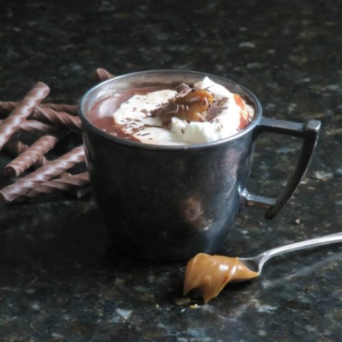 dark gray ceramic mug holding dulce de leche hot chocolate with drambuie and topped with whipped cream and chocolate shavings sits on a black marble surface with a silver spoon holding dulce de leche in front and chocolate sticks in the background