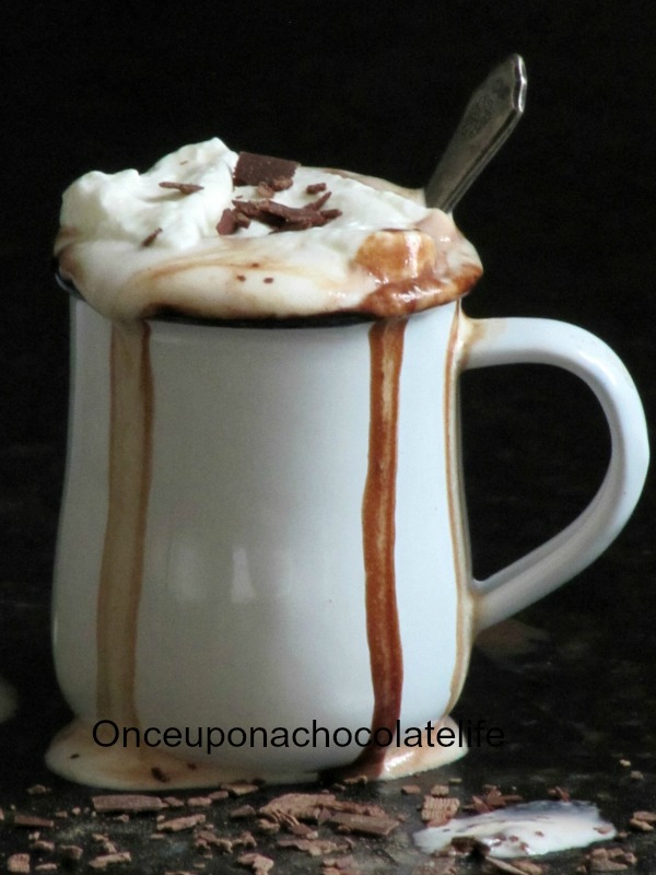 a large white handled mug holds hot chocolate made with fleur de sel and topped with whipped cream and chocolate shavings, with hot chocolate dripping down the sides of the mug. The mug is on a black marble surface surrounded by chocolate shavings against a dark background.