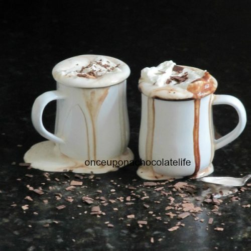 Two tall white mugs of fleur de sel hot cocoa with whipped cream and chocolate shavings topping and hot chocolate spilling over the tops and dripping down the sides are on a black surface that is covered in chocolate shavings against a black background.