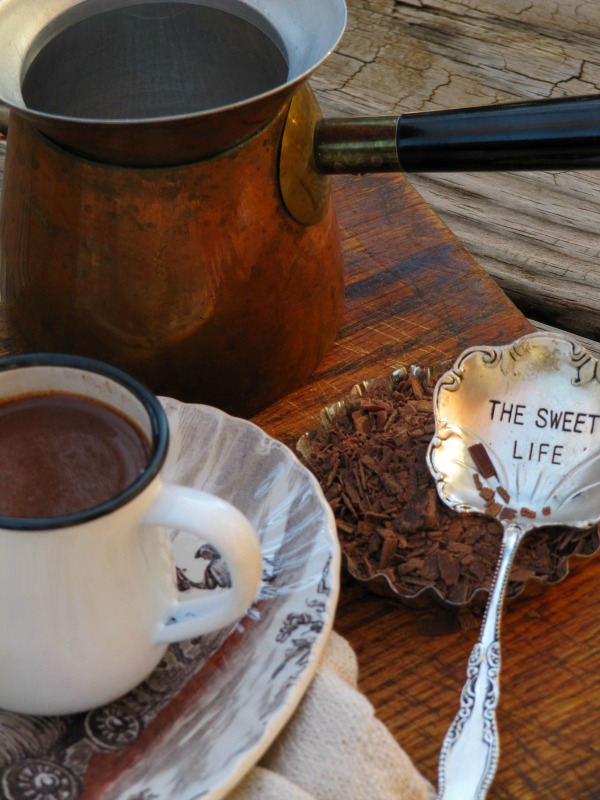 closeup of a white mug with blue rim filled with hot cocoa on atop view of two white enamel mugs with hot cocoa on a blue and white vintageoval platter, sitting on a white tray. The tray is on a wood table alongside a silver vintage spoon that says "The Sweet Life" and a vintage coffee ibrik with white ceramic mouth and blue enamel handle.