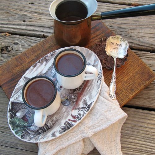 top view of two white enamel mugs with hot cocoa on a blue and white vintageoval platter, sitting on a white tray. The tray is on a wood table alongside a silver vintage spoon athat says "Sweet Life" and a vintage copper coffee pot with white ceramic mouth and blue bakelite handle.