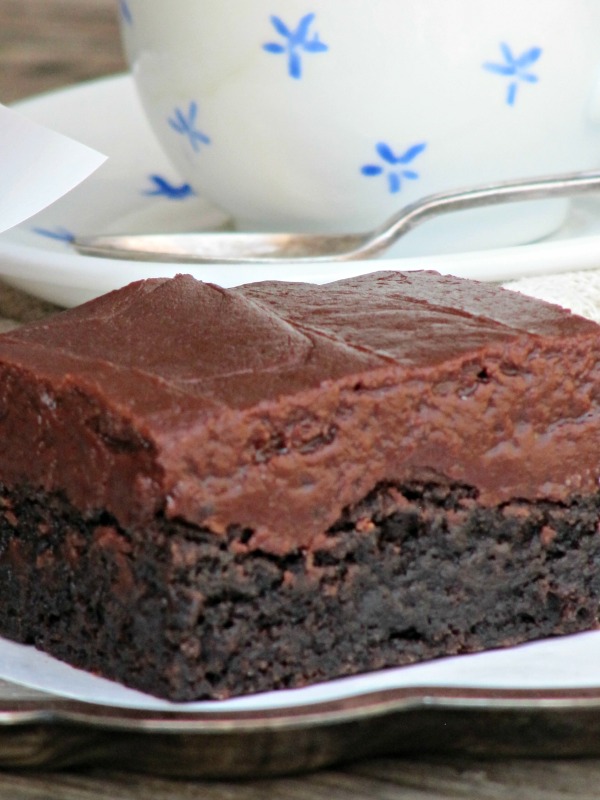 Close up of a rich thick chocolate fudge brownie with fudge frosting in front of a blue and white coffee cup on a white saucer.