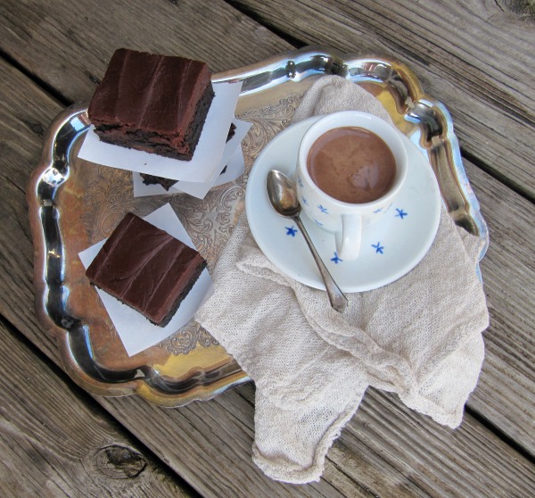 Overhead view of two fudge brownieswith fudge frosting on a serving platter on a wooden surface along with a blue and white cup of coffee on a white saucer.
