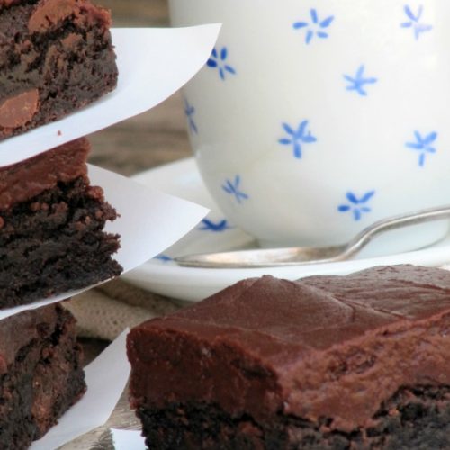 Dark chocolate brownie with chocolate fudge frosting on a plate next to a stack of three more brownies srparated by pieces of white parchment paper. The brownies are in front of a white mug decorated with blue decorations
