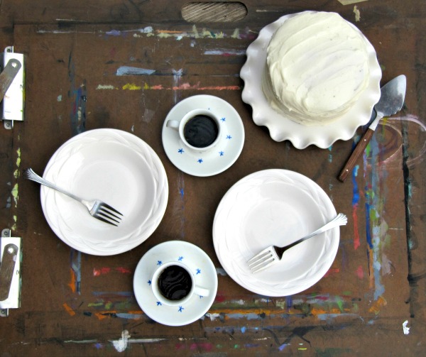 overhead view of a wooden surface with a white buttercream coated chocolate cake next to two plates with coffee.
