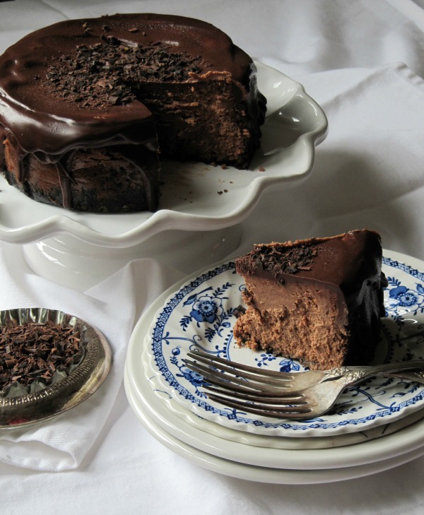 Slice of chocolate truffle cheesecake with ganache topping and chocolate curls next to a round chocolate cheesecake and a bowl of chopped chocolate.