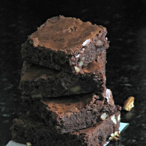 Stack of three chocolate brownies on a piece of white parchment paper cooling rack and pecan pieces scattered around on a gray table .