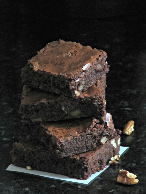 Stack of three chocolate brownies on a piece of white parchment paper cooling rack and pecan pieces scattered around on a gray table.