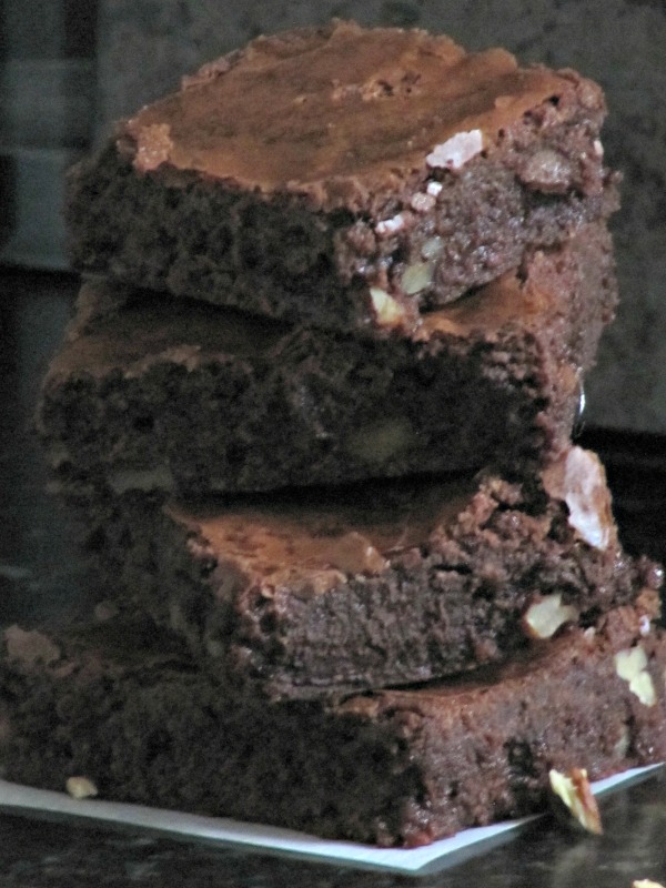 Close up of a stack of three chocolate brownies on a piece of white parchment paper cooling rack and pecan pieces scattered around on a gray table.