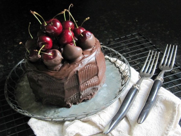 side view of chocolate cherry filled cake with chocolate covered cherries and whole fresh cherries on top set on a light gray platter on a white cloth on a black table with silver forks on the side.