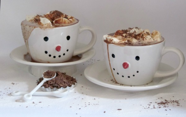 two round white mugs with smiley faces are filled with nutella hazelnut flavored hot cocoa and topped with whipped cream and chocolate shavings. The cups are next to a small white bowl of chopped dark chocolate pieces.