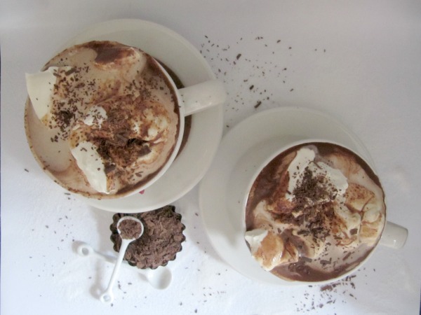 overhead view of two white mugs of hazelnut hot chocolate with whipped cream topping and shavings of chocolate sitting next to a small bowl of chopped chocolate.