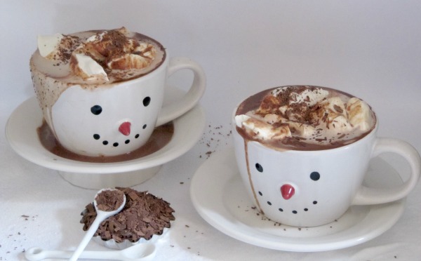Two white smiley-face mugs full of Nutella hot chocolate topped with whipped cream and chocolate shavigs are on white saucers on a white surface next to a small white bowl of shaved chocolate with a spoon in it