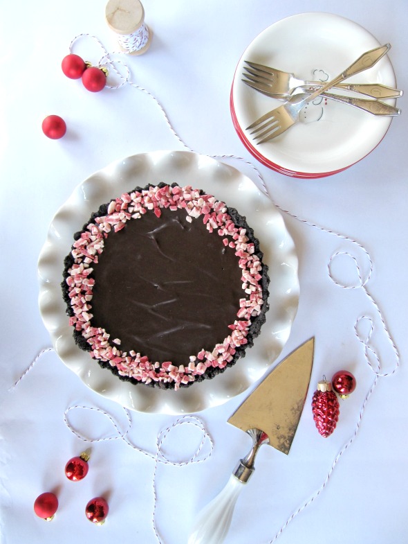 top view of a no bake chocolate peppermint tart ringed with crushed peppermint candy on a white platter on a table with red decorative balls and a stack of pink-rimmed dessert plates and dessert forks on top of the plates along with a silver pie server to the side of the tart..