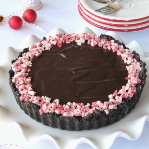 top view of round chocolate tart with a ring of crushed peppermint candy around the edge on a fluted white platter on a table with a stack of dessert plates and red ball decorations.