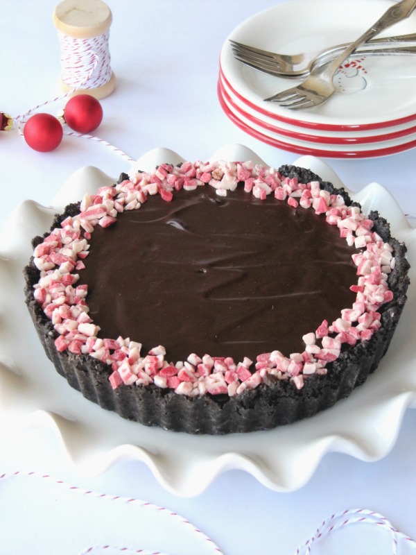 top view of round chocolate tart with a ring of crushed peppermint candy around the edge on a fluted white platter on a table with a stack of dessert plates and red ball decorations.