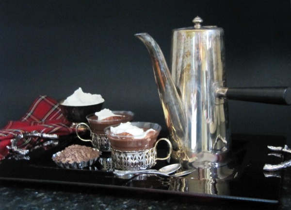 two antique silver cups filled with espresso hot chocolate with whipped cream topping are in front of a tall silver coffee pot and next to a bowl of shaved chocolate on a dark table against a dark background.