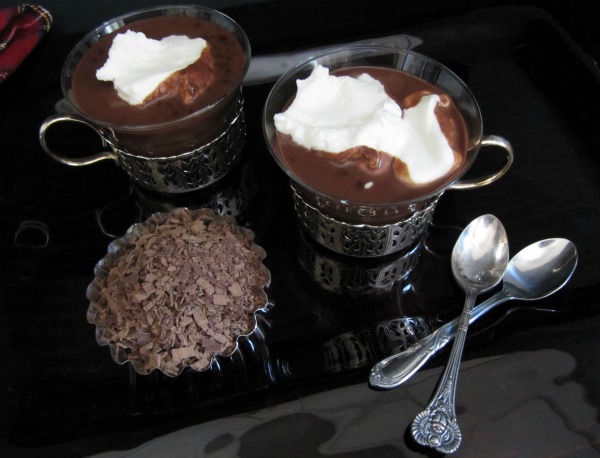 top view of two cups made of glass with silver cup holders filled with espresso hot chocolate drink topped with whipped cream. The cups are sitting on a dark table  next a bowl of chocolate shavings and two crossed silver spoons against a dark background.