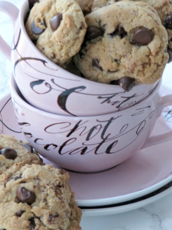 close up shot of a stack of two round pink cocoa mugs with writing on them on a pink saucer sitting on a white table top. The top mug is full of chocolate chip cookies. Two chocolate chip cookies are leaning against the saucer on the white table top.