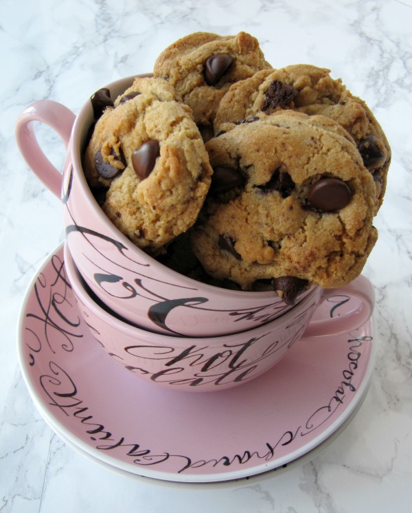 Top view of a stack of two round pink cocoa mugs with writing on them on a pink saucer. sitting on a white table top. The top mug is full of the best basic chocolate chip cookies.