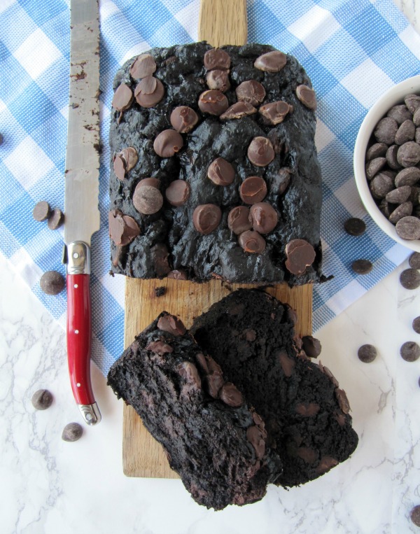 Top view of double chocolate banana bread loaf from Simtten Kitchen on a wood cutting board with two slices in front. Chocolate chips are on top of the loaf and scattered around the loaf,  a white bowl full of chocolate chips to the right and a red handled knife to the left of the loaf. 