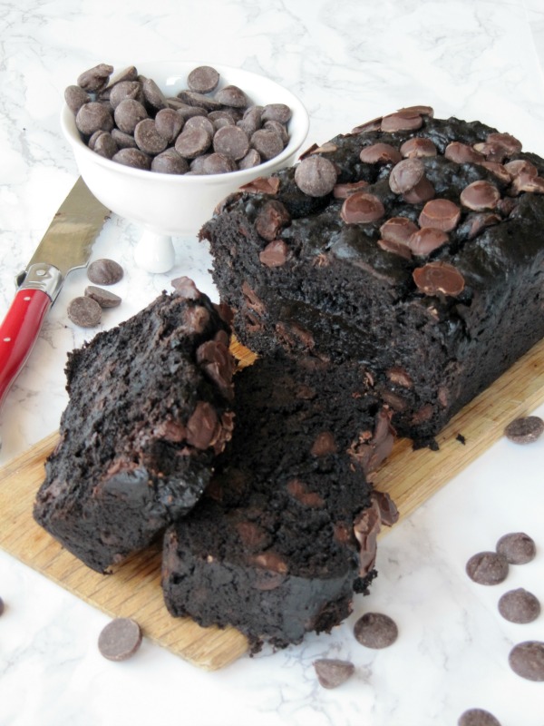 Image of double chocolate banana bread cut loaf with chocolate chips on top and two slices in front on a wood cutting board with a white bowl of chocolate chips to the side and chocolate chips scattered around the board. Board is on a white table and a red handled knife is to the left side of the loaf.