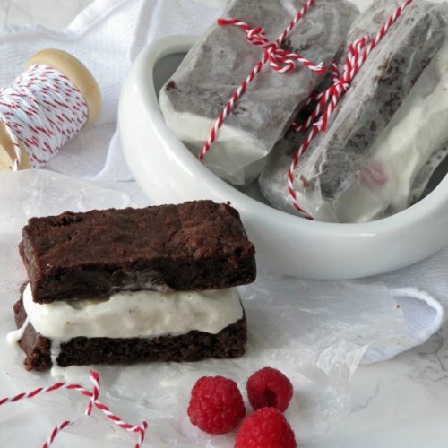 A large rectangulaar brownie ice cream sandwich with vanilla raspberry ice cream filling is sitting on a white surface in front of a bowl of wrapped ice cream sandwiches and has three fresh raspberries in front of it on the table that also has a roll of red twine sitting on it