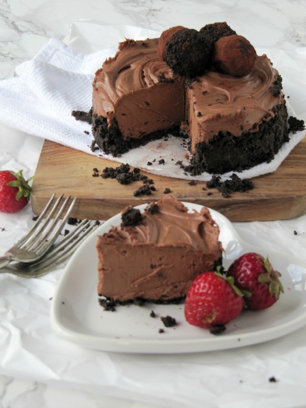 Round no bake chocolate cheesecake with oreo crust and decorated with chocolate truffles sits on a cutting board behind a large slice of the cake on a heart-shaped white plate garnished with fresh strawberries