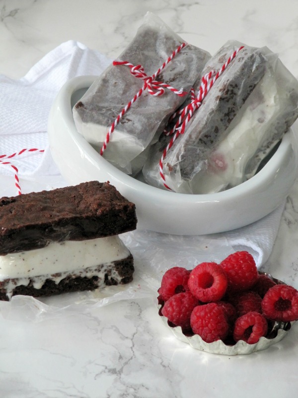 A large brownie raspberry ice cream sandwich filled with homemade raspberry vanilla ice cream is sitting on a table next to a bowl of fresh red raspberries, a white ceramic bowl with two ice cream sandwiches wrapped in white parchment paper and red and white twine. 