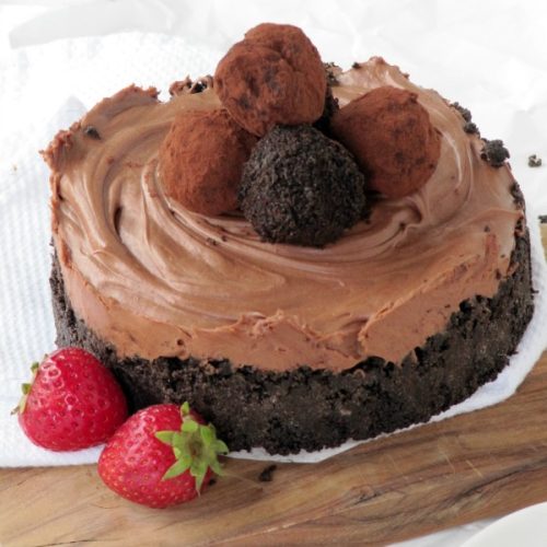 large round chocolate cheesecake with a dark chocolate Oreo crust and decorated with homemade chocolate truffles on top sits on a wood cutting board along with two fresh strawberries as a garnish.