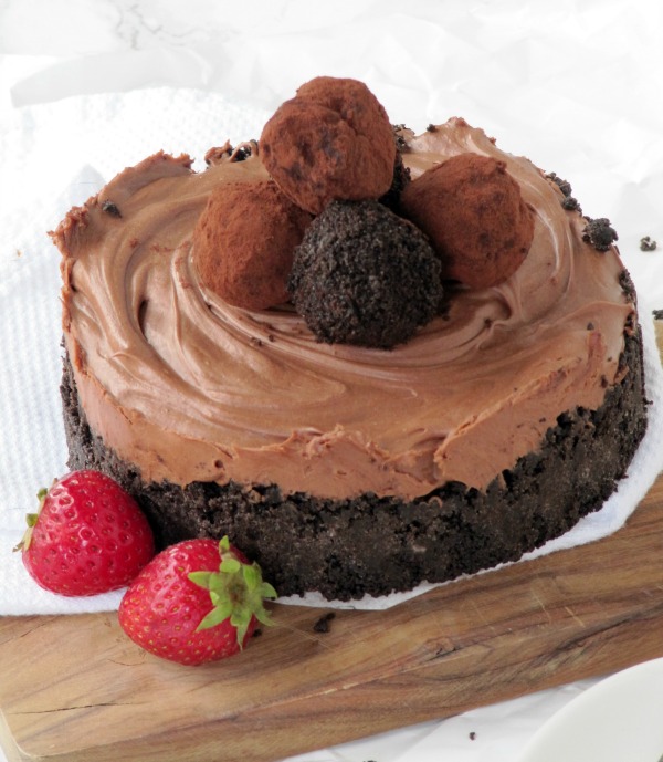 large round no bake chocolate cheesecake chocolate truffles on top sits on a wood cutting board along with two fresh strawberries as a garnish.