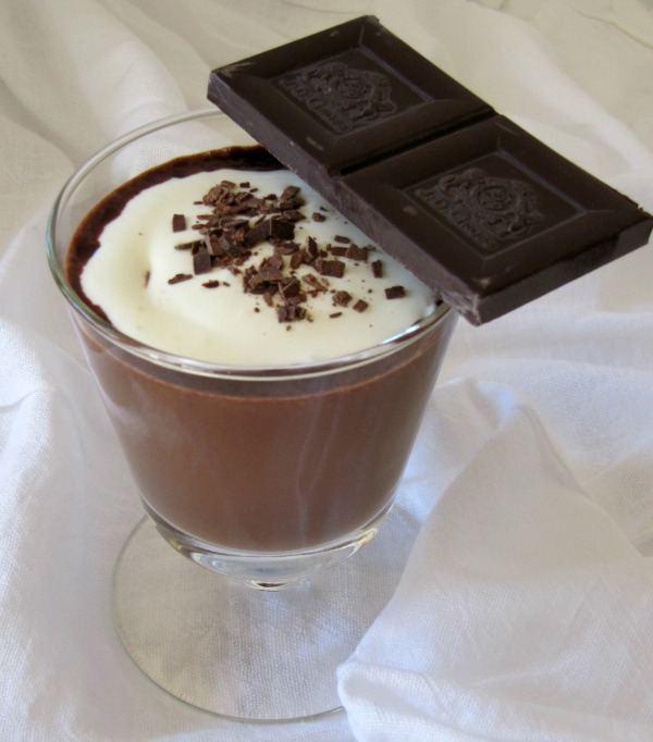 Side view of a glass cup of dark chocolate atole beverage made with cocoa powder, dark chocolate, milk, sugar and corn meal, with a large piece of dark chocolate sitting on the rim of the cup.