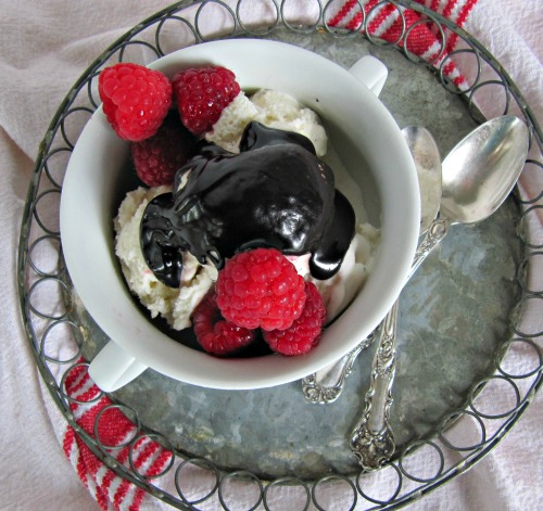 closeup overhead view of a silver serving tray holding two silver spoons and a white ceramic bowl of ice cream drenched with bailey's hot fudge sauce and several fresh raspberries.