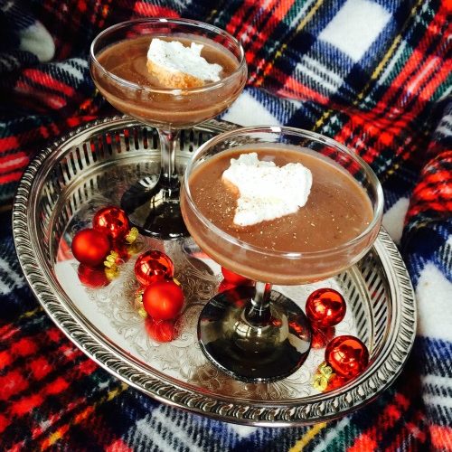 Two footed glasses on an oval silver tray are filled with frozen hot chocolate bailey's cocktail. The tray is on a red plaid covtable covering and round red christmas ornaments are on the tray