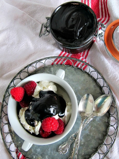 overhead view of a round metal serving tray holding vanilla ice cream covered with bailey's hot fudge sauce and garnished with fresh raspberries. The tray is on a red and white table cloth with a jar of hot fudge sauce nearby.