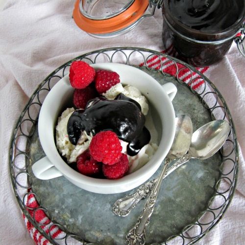overhead view of a white tablecloth with a red stripe on it holding a round metal serving tray with two silver spoons and a hot fudge sundae made with bailey's irish cream and dark chocolate garnished with fresh raspberries . A jar of hot fudge sauce is in the background.