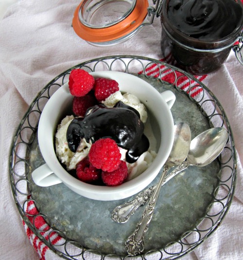 overhead view of a white tablecloth with a red stripe on it holding a round metal serving tray with two silver spoons and a hot fudge sundae made with bailey's hot fudge sauce with irish cream and dark chocolate garnished with fresh raspberries . A jar of hot fudge sauce is in the background.