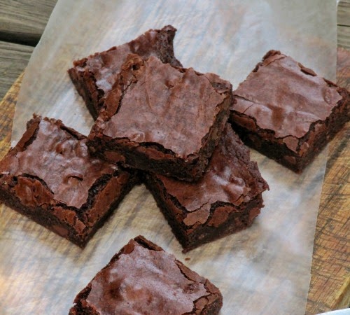 overhead view of six rich baked chocolate fudge brownies with crinkly tops sit on a piece of parchment paper on a wood surface.